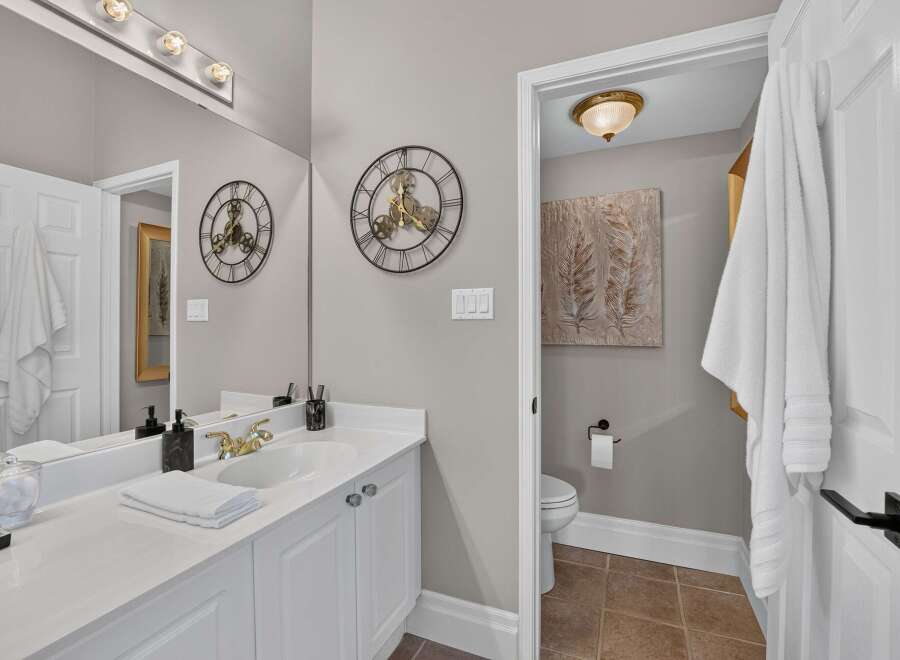 This elegant bathroom features a spacious vanity with a white countertop and gold-accented fixtures, complemented by a large mirror reflecting a stylish wall clock.