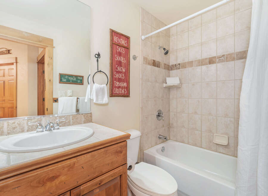 This charming bathroom features a spacious vanity with a white sink and chrome fixtures, complemented by a large mirror reflecting the wooden door and a decorative sign.