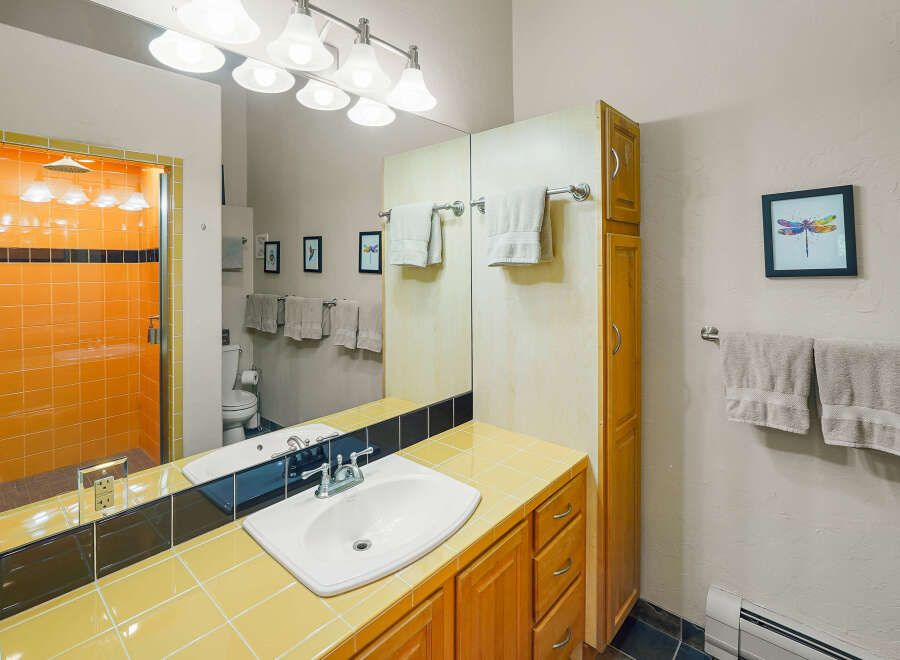 This vibrant bathroom features a striking orange tiled shower with a glass door, complemented by a sunny yellow tiled countertop and backsplash.