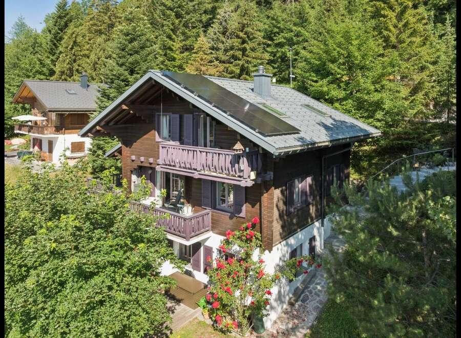 Charming wooden chalet nestled amidst lush green trees, featuring multiple balconies with traditional railings and purple shutters.