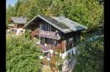 Charming wooden chalet nestled amidst lush green trees, featuring multiple balconies with traditional railings and purple shutters.