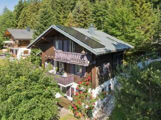 Charming wooden chalet nestled amidst lush green trees, featuring multiple balconies with traditional railings and purple shutters.