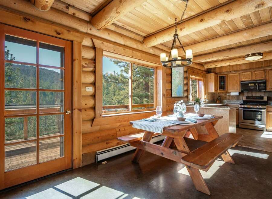 Experience rustic charm in this log cabin dining area, featuring a wooden picnic table set for a meal with stunning mountain views through large windows.
