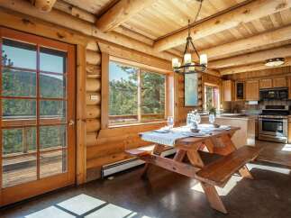 Experience rustic charm in this log cabin dining area, featuring a wooden picnic table set for a meal with stunning mountain views through large windows.