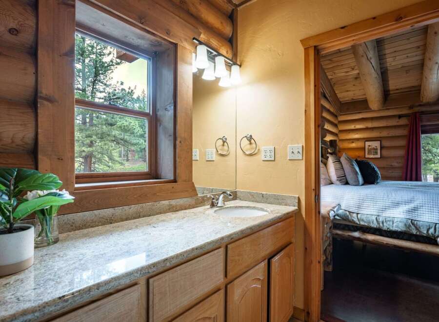 Experience rustic charm in this log cabin bathroom featuring a granite-topped vanity and a large window with picturesque forest views.