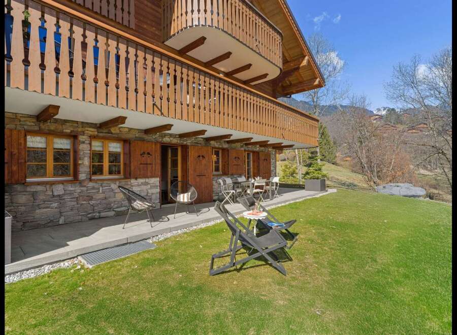 Experience alpine living in this charming wooden chalet featuring a stone facade and a spacious balcony overlooking a lush green lawn.
