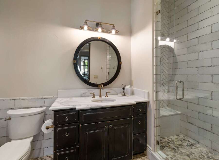 This charming bathroom features a dark wood vanity with a marble countertop, complemented by a round black framed mirror and a three-light vanity light fixture.