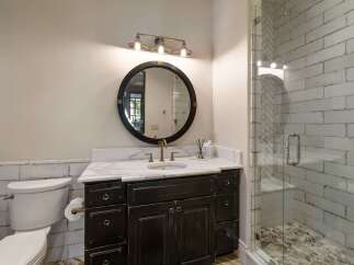 This charming bathroom features a dark wood vanity with a marble countertop, complemented by a round black framed mirror and a three-light vanity light fixture.