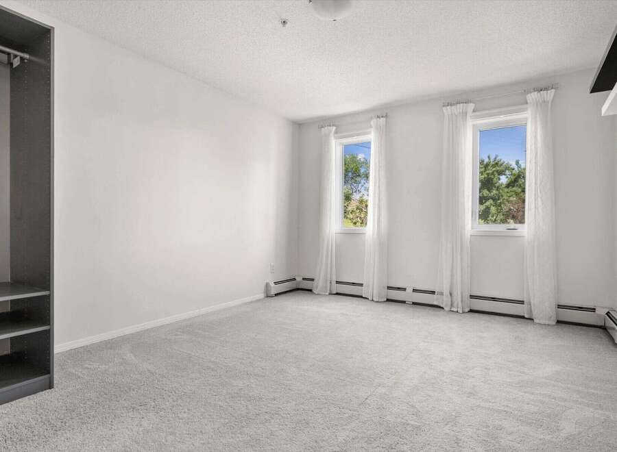 This spacious bedroom features a large, open layout with plush grey carpeting and ample natural light streaming through two large windows.