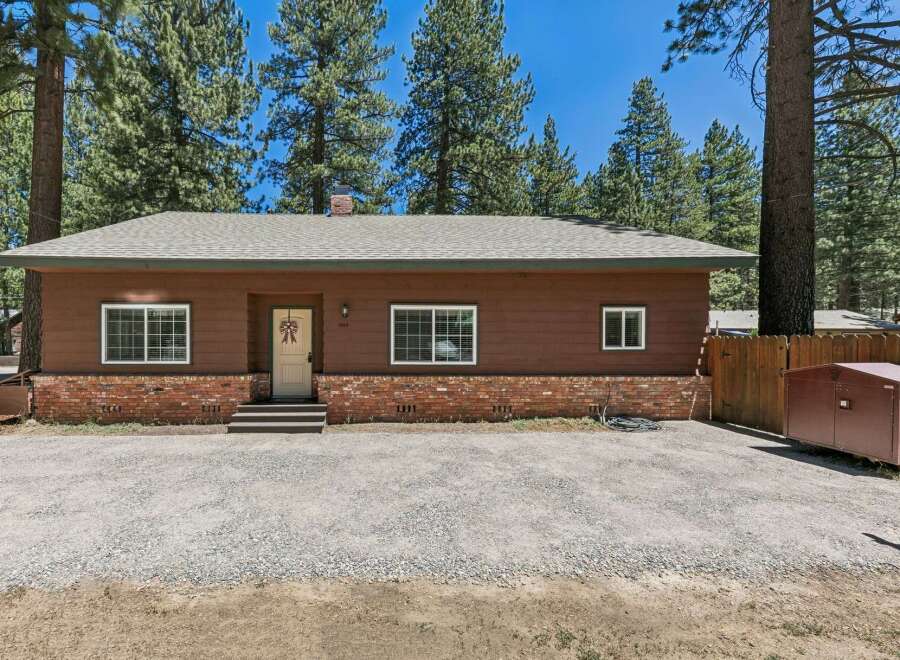 Charming single-story home nestled among tall pine trees, featuring a warm brown exterior with a classic brick foundation.