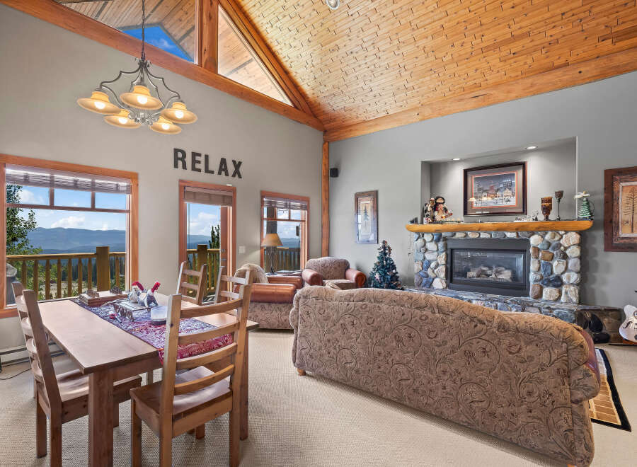 Experience breathtaking mountain views from this stunning living and dining area, featuring a cozy stone fireplace and a rustic wooden ceiling.