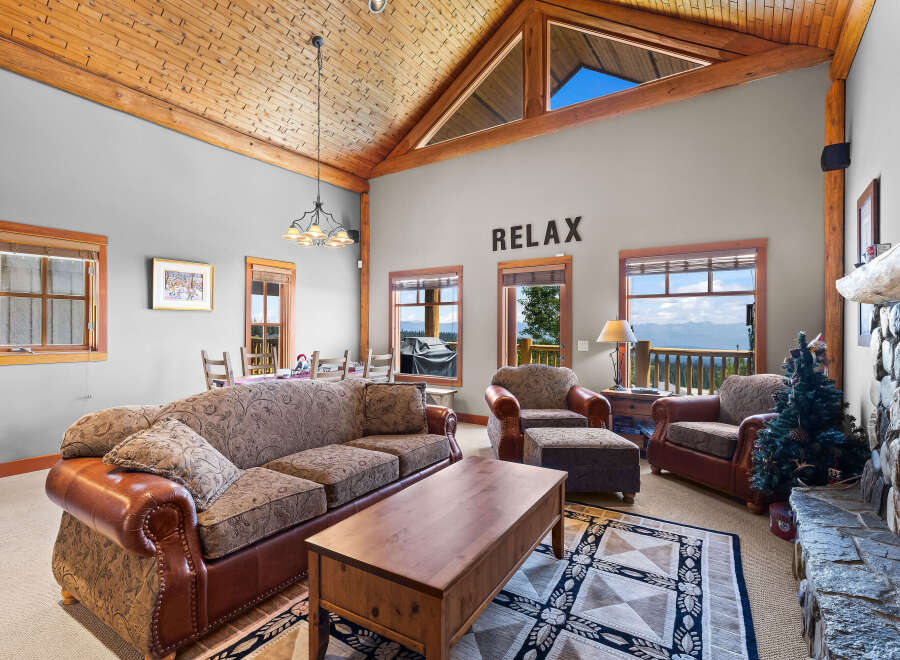 Experience breathtaking mountain views from this stunning living room, featuring a vaulted wood-paneled ceiling and a cozy stone fireplace.