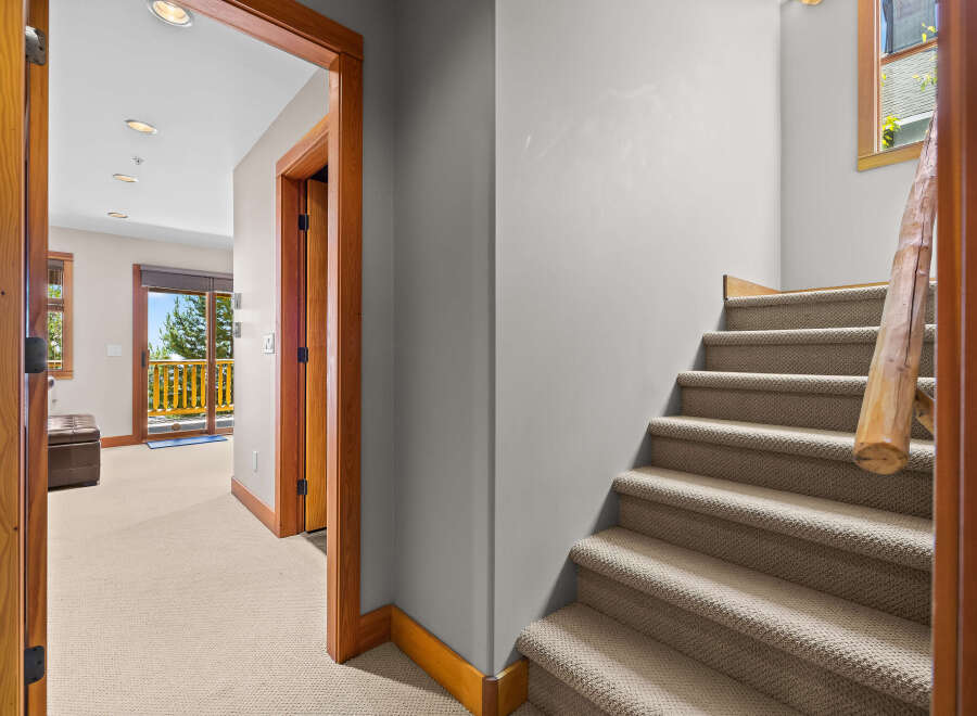 Step into this inviting space featuring a carpeted staircase with a rustic wooden handrail, leading to a bright hallway with a leather ottoman and a sliding glass door to a balcony.