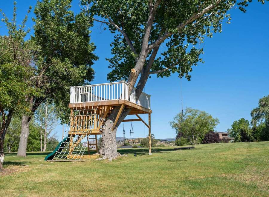 Experience outdoor fun with this incredible treehouse featuring a slide, climbing net, and swings, all set against a backdrop of lush greenery and a clear blue sky.
