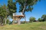 Experience outdoor fun with this incredible treehouse featuring a slide, climbing net, and swings, all set against a backdrop of lush greenery and a clear blue sky.