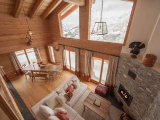 Experience luxury mountain living in this stunning chalet, featuring a spacious open-plan living area with a cozy stone fireplace and large windows offering breathtaking snowy mountain views.