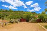 Discover this unique property featuring rustic red barns nestled against a lush, green hillside under a vibrant blue sky with fluffy white clouds.