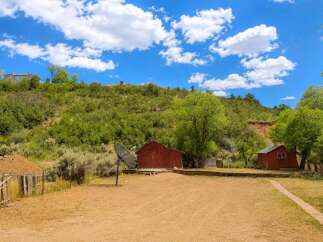 Discover this unique property featuring rustic red barns nestled against a lush, green hillside under a vibrant blue sky with fluffy white clouds.
