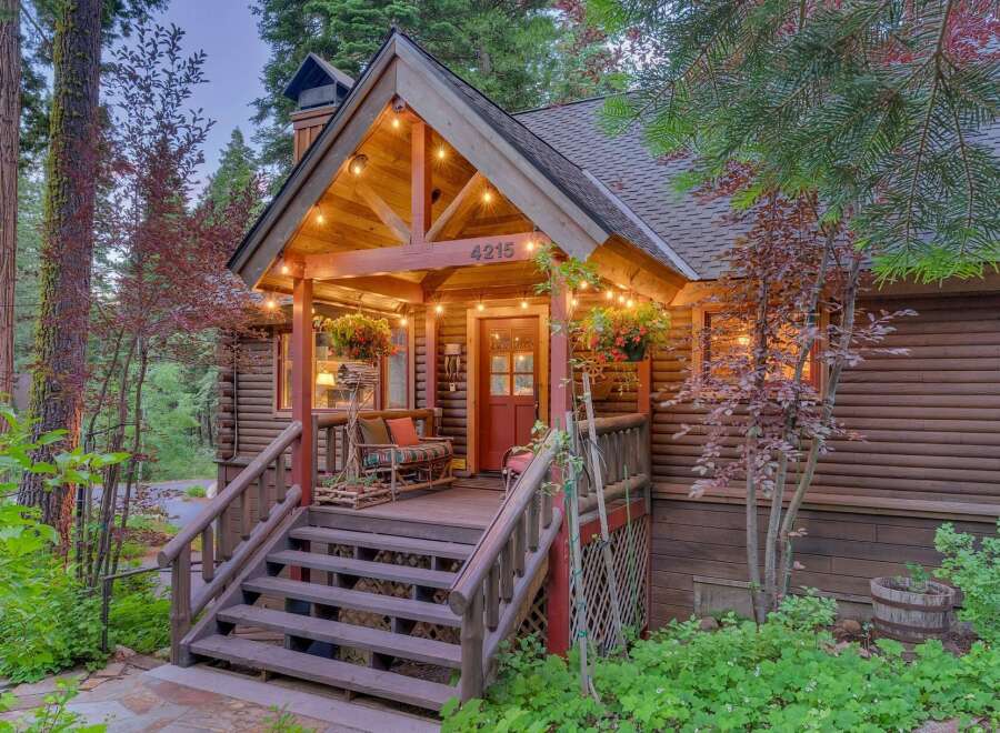 Charming log cabin nestled amongst tall trees, featuring a welcoming porch with string lights and a cozy seating area.