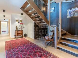 Experience luxury living in this stunning home featuring a modern wooden staircase with glass railings, complemented by a vibrant Persian rug and elegant wall sconces.