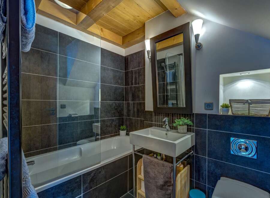 This modern bathroom features a sleek bathtub with a glass enclosure, complemented by dark tiled walls and a wooden ceiling.