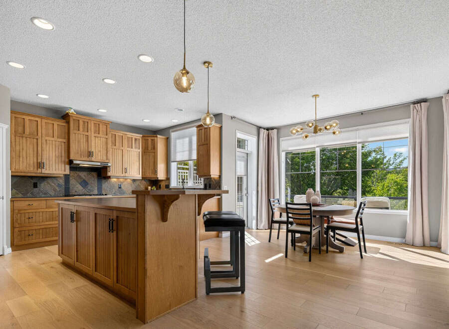 This spacious kitchen and dining area boasts beautiful natural light streaming through large windows, highlighting the warm wood cabinetry and elegant pendant lighting.