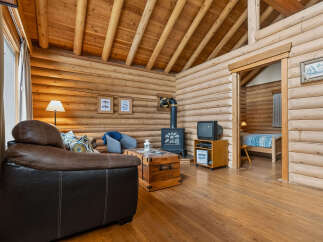 Experience rustic charm in this cozy log cabin living space, featuring a comfortable leather sofa, a wood-burning stove for warmth, and a charming wooden trunk coffee table.