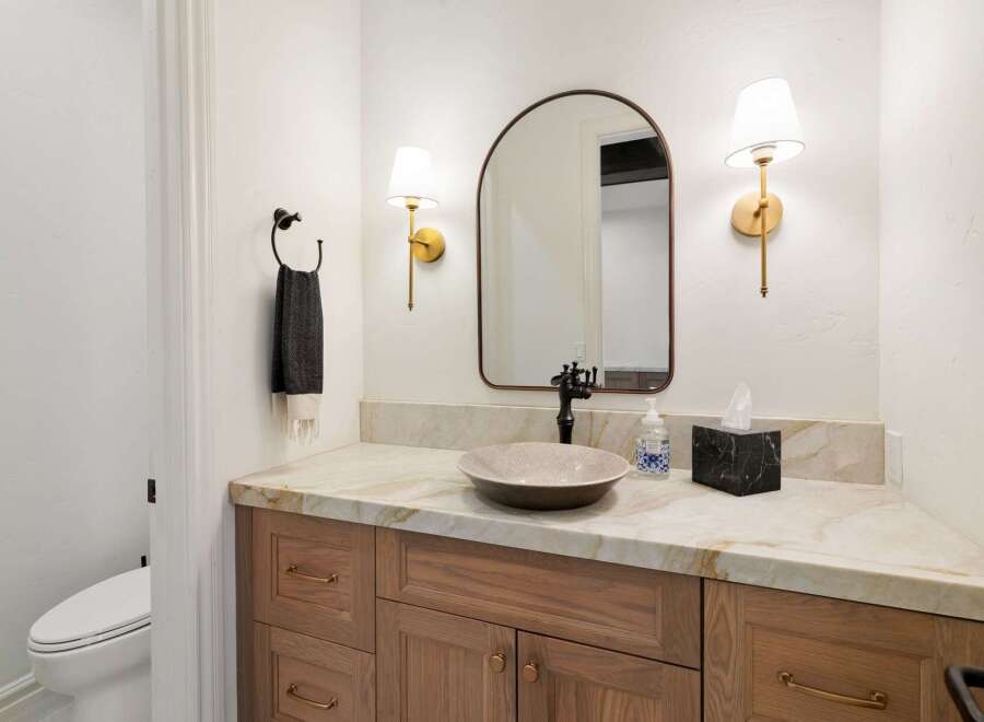 This modern bathroom features a stylish vanity with a stone countertop and a vessel sink, complemented by a large arched mirror and elegant brass sconces.