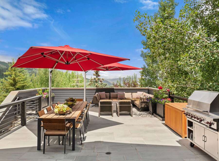 Enjoy al fresco dining and entertaining on this expansive deck, featuring a large wooden dining table with chairs and two vibrant red umbrellas for shade.
