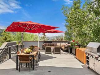 Enjoy al fresco dining and entertaining on this expansive deck, featuring a large wooden dining table with chairs and two vibrant red umbrellas for shade.