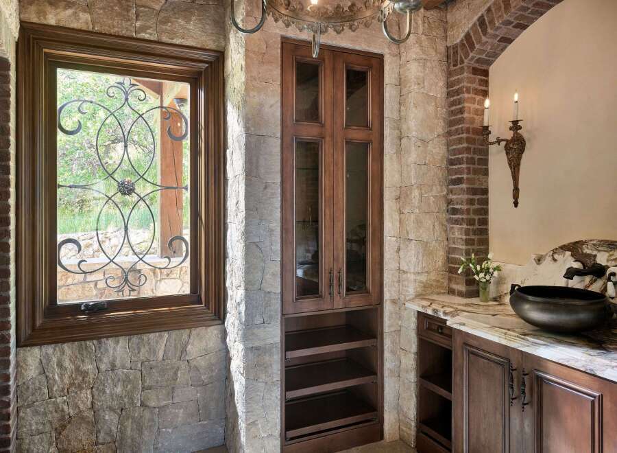 This rustic bathroom features a large stone fireplace, a dark wood vanity with a marble countertop, and a unique vessel sink.