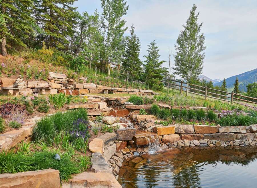 Discover this stunning mountain property featuring a serene tiered water feature cascading into a tranquil pond, surrounded by lush landscaping and natural stone accents.