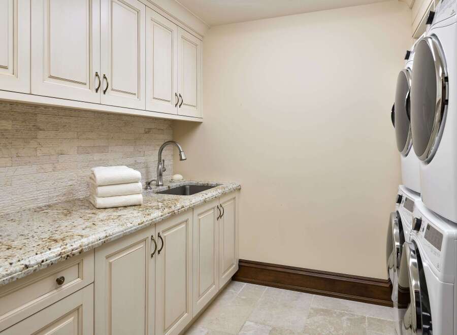 This laundry room features elegant cream-colored cabinetry with ornate hardware, complemented by a speckled granite countertop and a stacked stone backsplash.