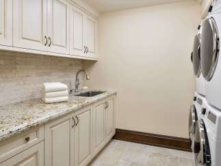This laundry room features elegant cream-colored cabinetry with ornate hardware, complemented by a speckled granite countertop and a stacked stone backsplash.