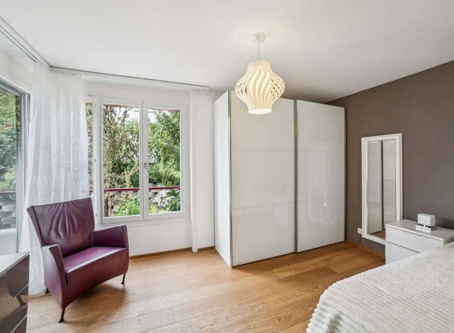 Discover this inviting bedroom featuring a comfortable purple armchair by the large window, offering ample natural light and a view of lush greenery.