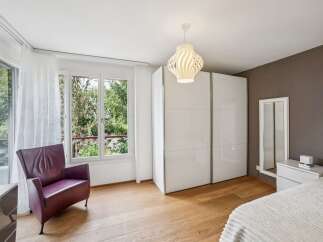 Discover this inviting bedroom featuring a comfortable purple armchair by the large window, offering ample natural light and a view of lush greenery.
