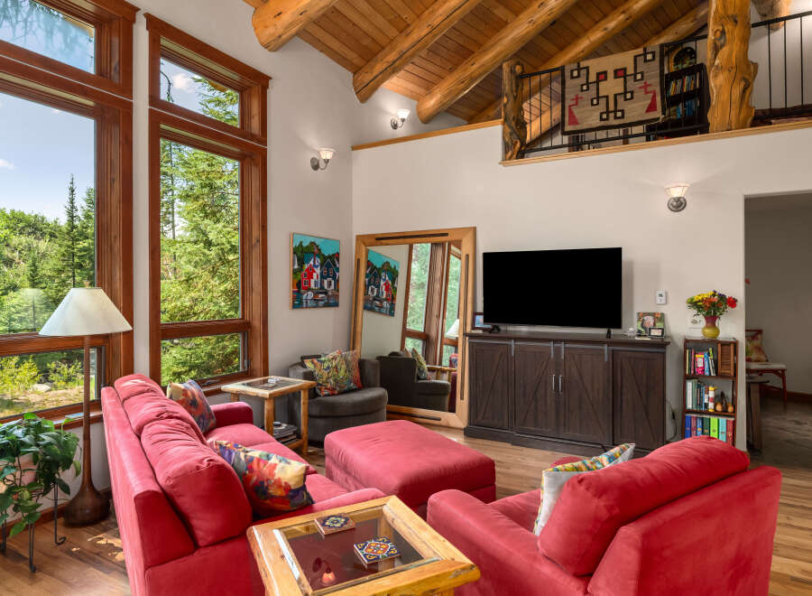 Experience rustic charm in this spacious living room featuring a vaulted wood-beamed ceiling and large windows offering serene forest views.