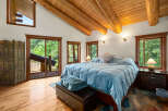 Experience rustic charm in this spacious bedroom featuring a vaulted wood-beamed ceiling and large windows offering serene forest views.