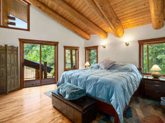 Experience rustic charm in this spacious bedroom featuring a vaulted wood-beamed ceiling and large windows offering serene forest views.