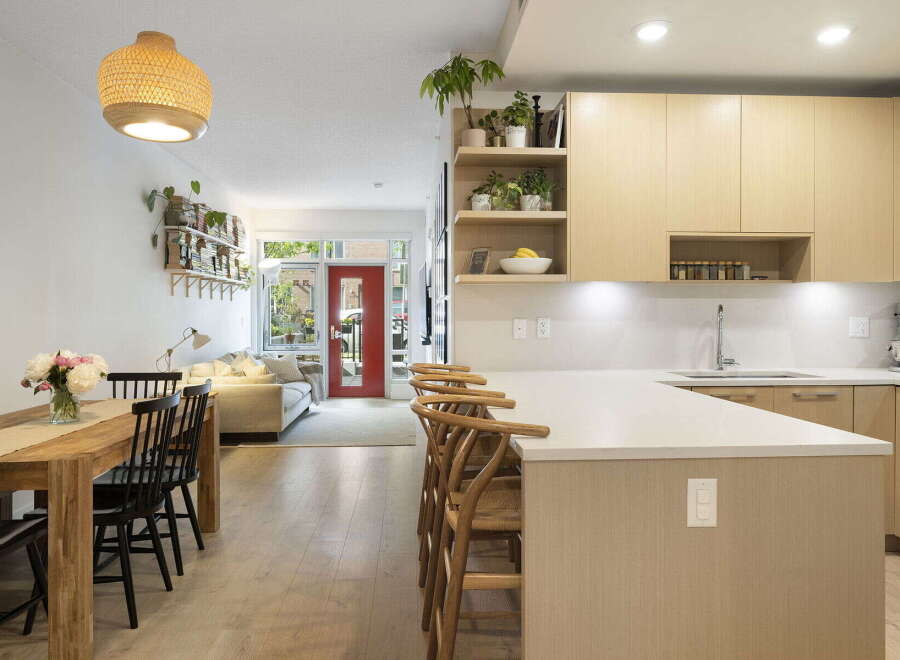 This modern open-concept living space features a stylish dining area with a wooden table and black chairs, flowing into a bright living room with a comfortable sofa and a striking red door.