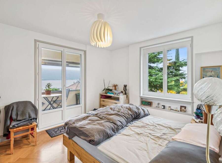 Experience serene living in this charming bedroom featuring a comfortable bed with a cozy duvet and a large window offering picturesque views of lush greenery and a distant lake.