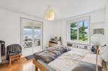 Experience serene living in this charming bedroom featuring a comfortable bed with a cozy duvet and a large window offering picturesque views of lush greenery and a distant lake.