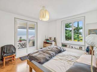 Experience serene living in this charming bedroom featuring a comfortable bed with a cozy duvet and a large window offering picturesque views of lush greenery and a distant lake.