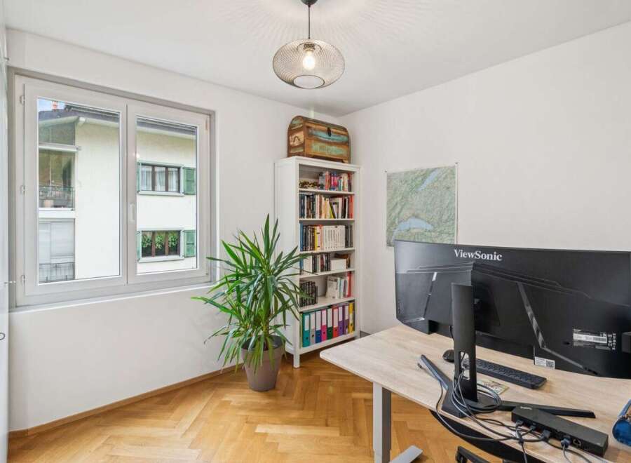 This home office features a large window offering natural light, a tall white bookshelf filled with books and binders, and a modern desk with dual monitors.