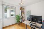 This home office features a large window offering natural light, a tall white bookshelf filled with books and binders, and a modern desk with dual monitors.