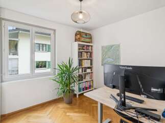 This home office features a large window offering natural light, a tall white bookshelf filled with books and binders, and a modern desk with dual monitors.