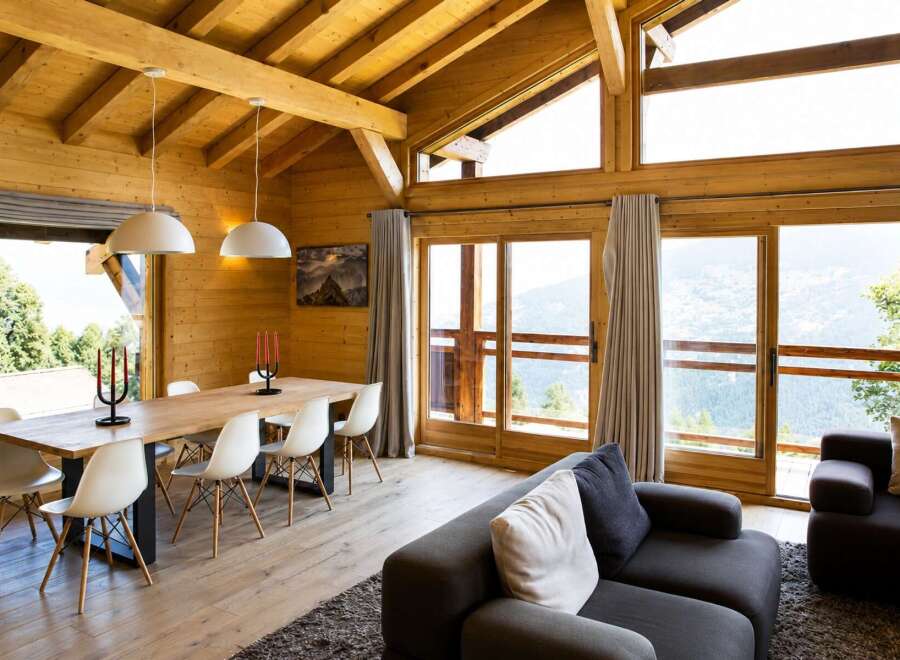 Experience breathtaking mountain views from this stunning wooden chalet, featuring a spacious dining area with a large wooden table and modern white chairs.
