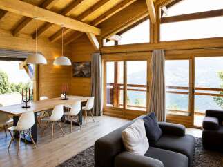 Experience breathtaking mountain views from this stunning wooden chalet, featuring a spacious dining area with a large wooden table and modern white chairs.