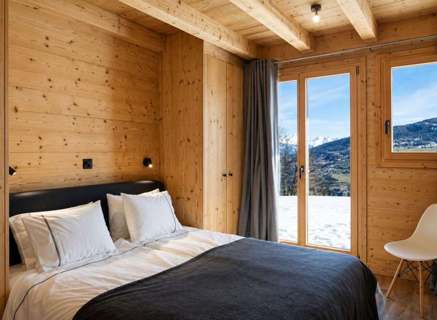 Experience cozy mountain living in this charming wooden bedroom, featuring a comfortable bed and stunning panoramic views of snow-capped peaks through large windows.