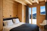 Experience cozy mountain living in this charming wooden bedroom, featuring a comfortable bed and stunning panoramic views of snow-capped peaks through large windows.